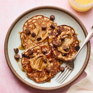 Plate with protein pancakes topped with maple syrup, peanut butter and chocolate chips. Glass or orange juice is next to the plate of pancakes.