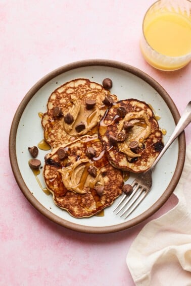 Plate with protein pancakes topped with maple syrup, peanut butter and chocolate chips. Glass or orange juice is next to the plate of pancakes.