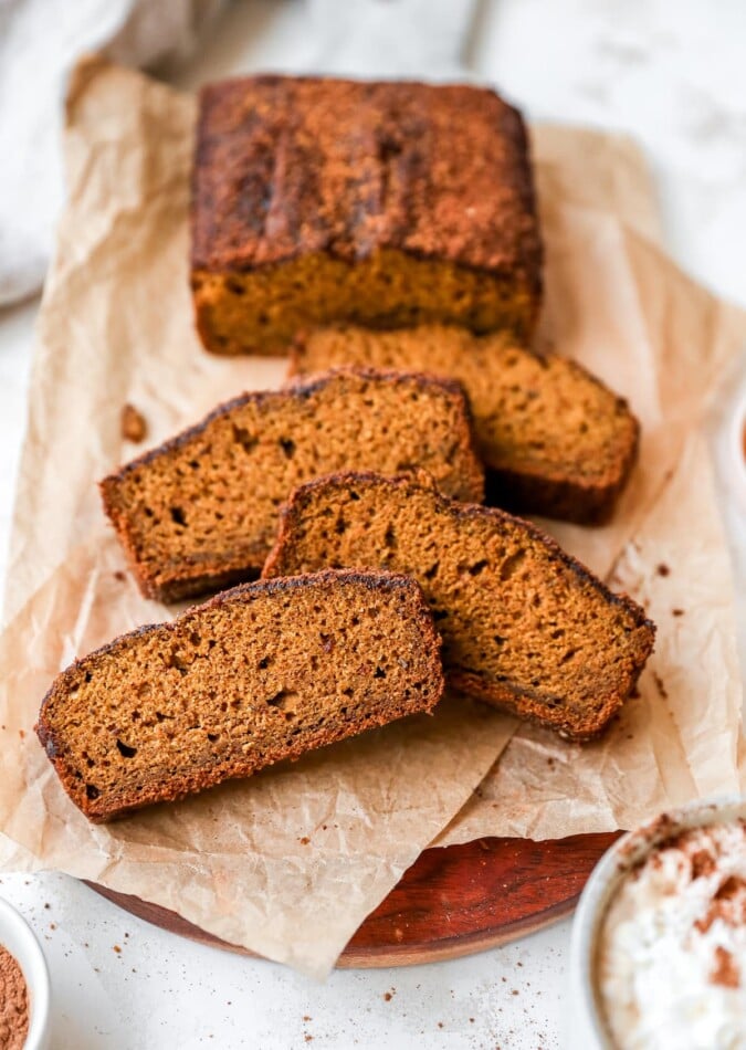 4 slices of pumpkin bread in focus with the remaining loaf in the background, resting on parchment paper on top of a cutting board.