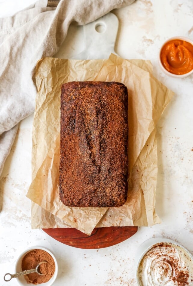 An overhead shot of a loaf of pumpkin bread, resting on parchment paper on top of a cutting board. Several ingredients in small dishes are scattered around the loaf of bread.