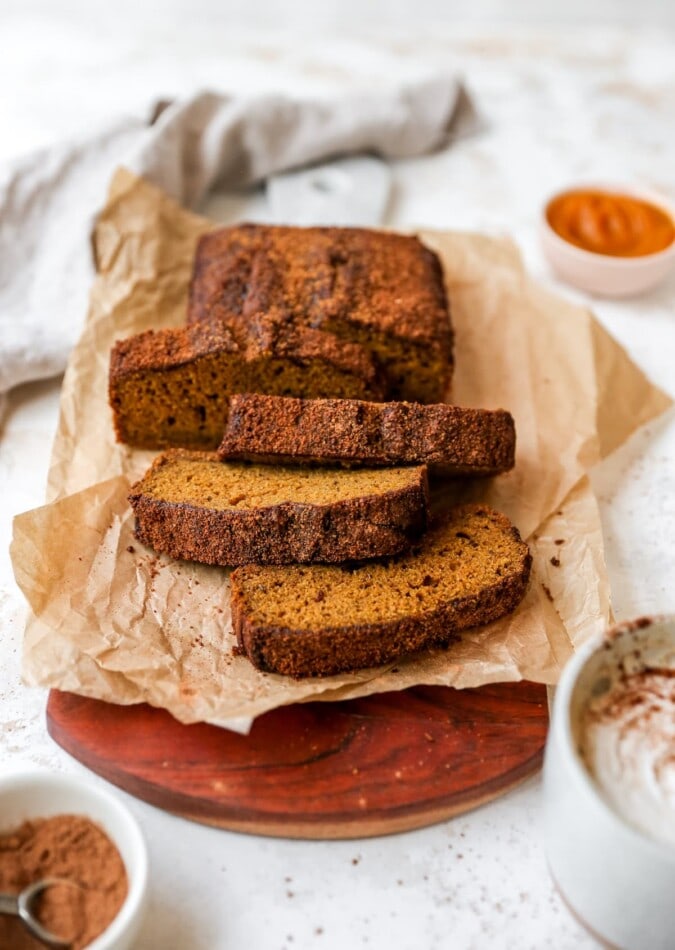 Loaf of pumpkin bread on a parchment paper resting on a cutting board. 4 slices have been cut from the loaf.