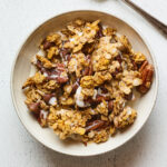 A bowl of pumpkin granola served in a bowl with milk. A silver spoon rests aside the bowl.