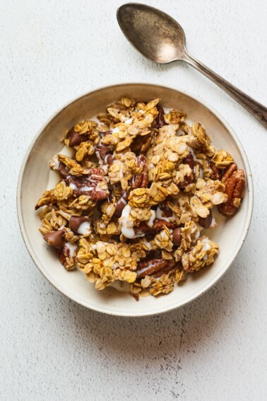 A bowl of pumpkin granola served in a bowl with milk. A silver spoon rests aside the bowl.