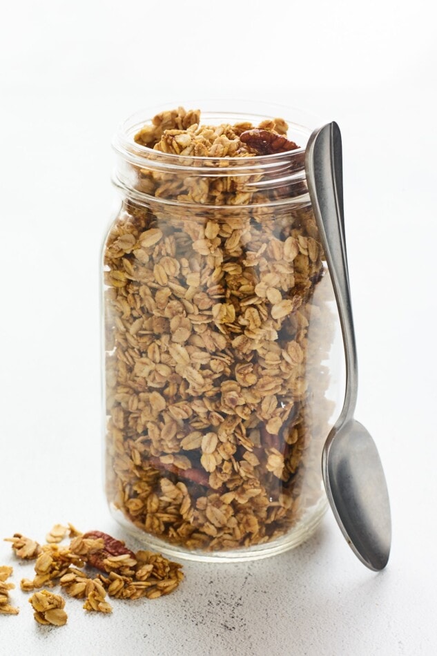 A mason jar filled with pumpkin granola. A silver spoon rests alongside the jar.