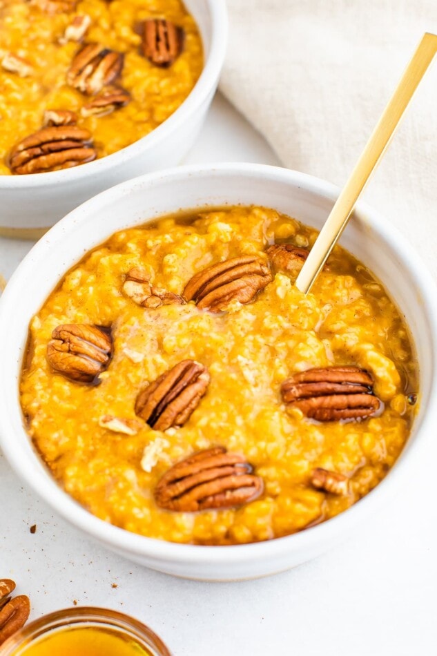Pumpkin oatmeal topped with whole pecans. A gold spoon rests inside the bowl.