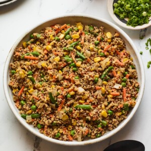 A serving bowl filled with quinoa fried rice. A black spoon rests below the bowl. Just out of frame is a bottle of sriracha and a bowl of green onions.