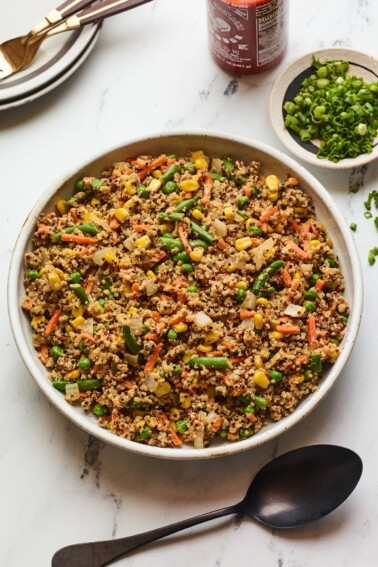 A serving bowl filled with quinoa fried rice. A black spoon rests below the bowl. Just out of frame is a bottle of sriracha and a bowl of green onions.