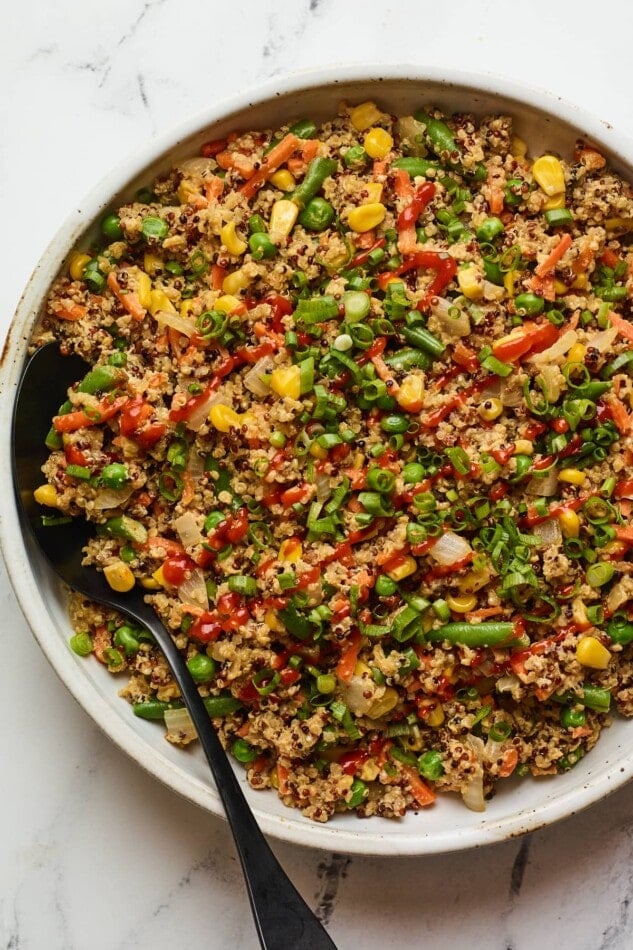 An overhead shot of a bowl of quinoa fried rice. Sriracha has been drizzled on top and a black serving spoon rests inside the bowl.