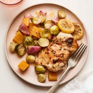 A plate with roasted chicken, lemon slices, Brussels sprouts, butternut squash, and onion. A silver fork rests on the plate.