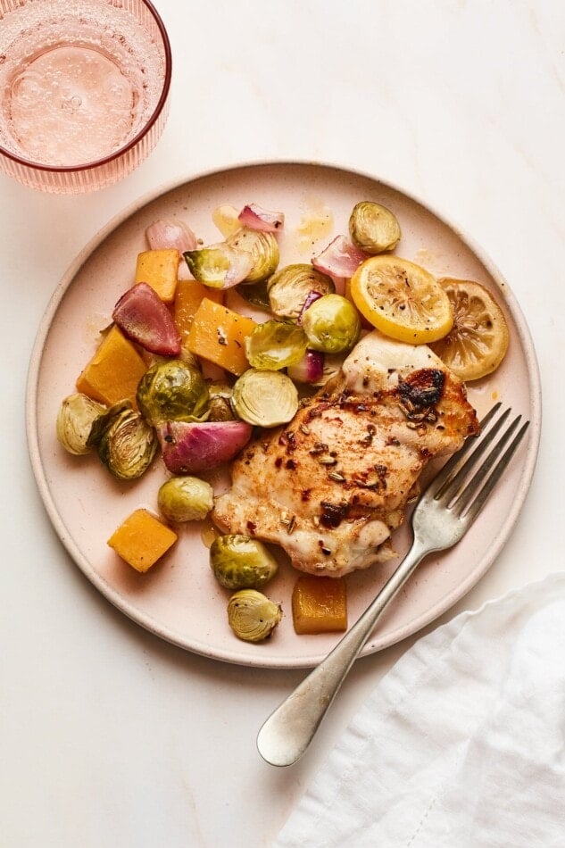 A plate with roasted chicken, lemon slices, Brussels sprouts, butternut squash, and onion. A silver fork rests on the plate.