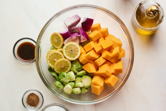 A glass bowl filled with butternut squash, onion, lemon slices, and Brussels sprouts. Additional ingredients are around the bowl.