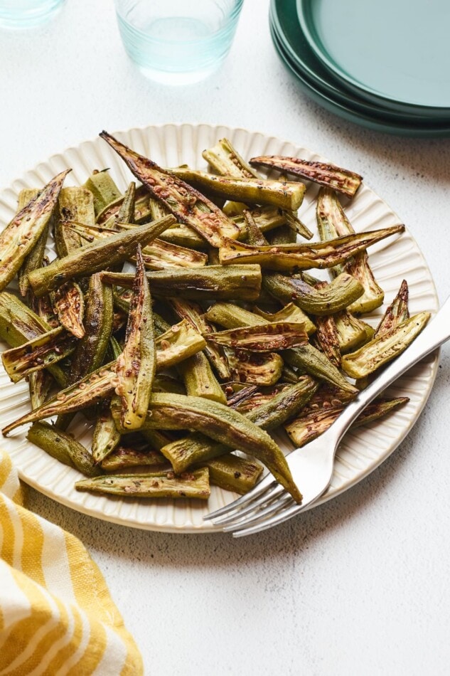 Roasted okra on a plate with a fork. A couple glasses of water and a stack of plates are beside the plate of okra.