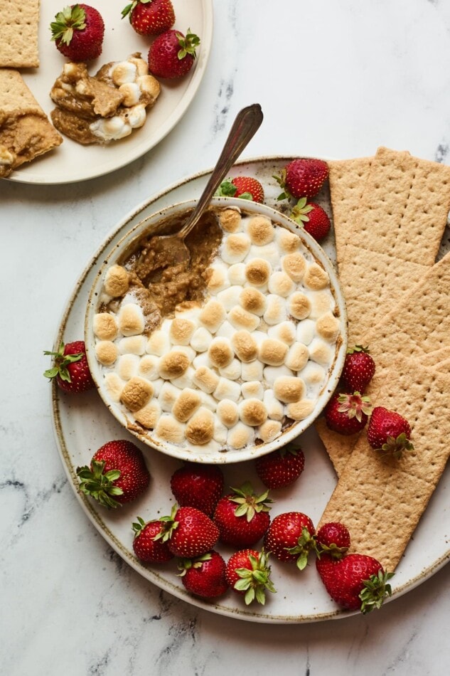 S'mores dip plated with strawberries and graham crackers for dipping. A spoon is taking a scoop from the dip.