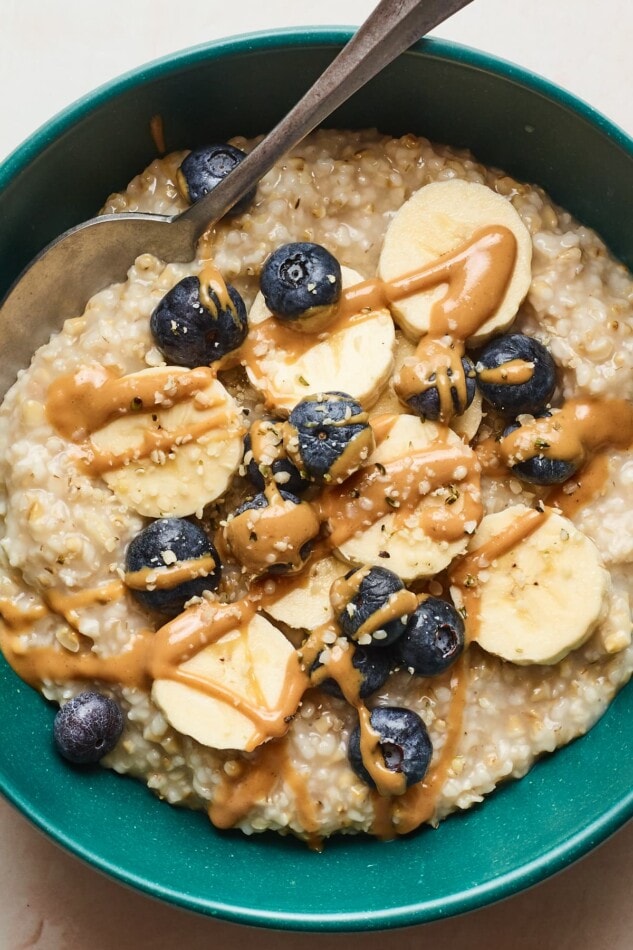 A close up shot of steel cut oats in a bowl with blueberries, bananas, and almond butter drizzled on top. A spoon rests in the bowl.