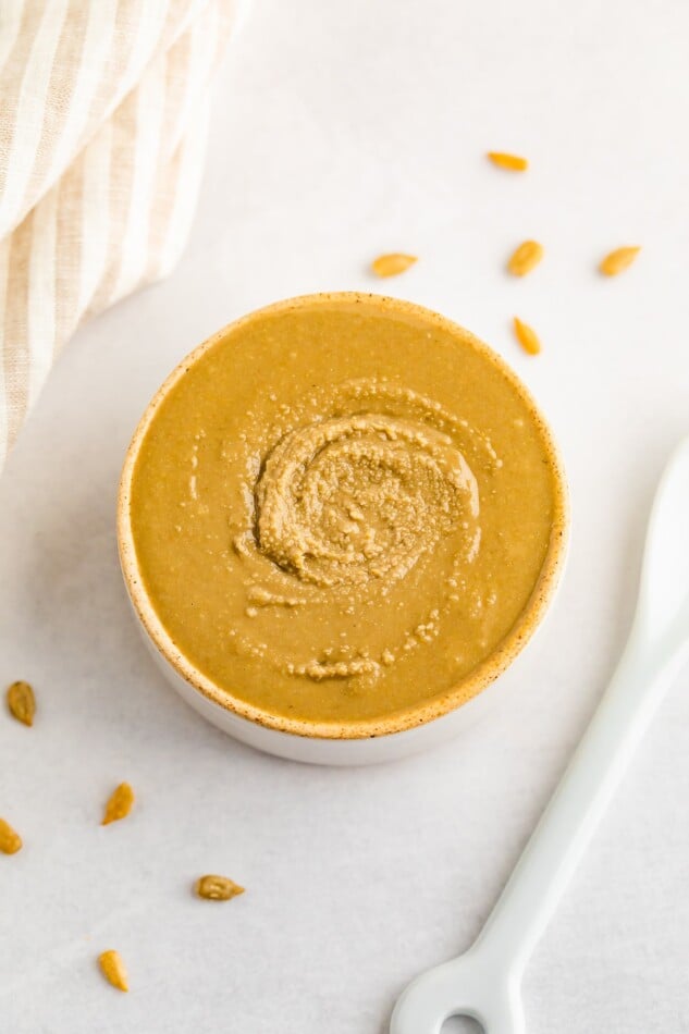 Overhead shot of sunflower butter in an open container with a spoon and some sunflower seeds scattered around.