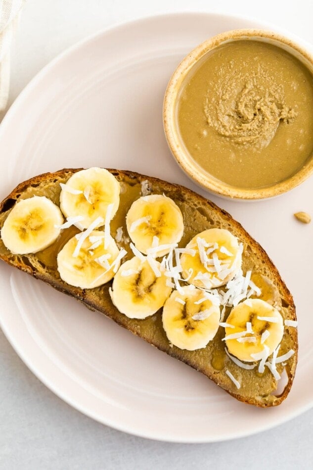 A plate with a piece of toast with sunflower seed butter, banana, and coconut flakes and a bowl of sunflower seed butter..