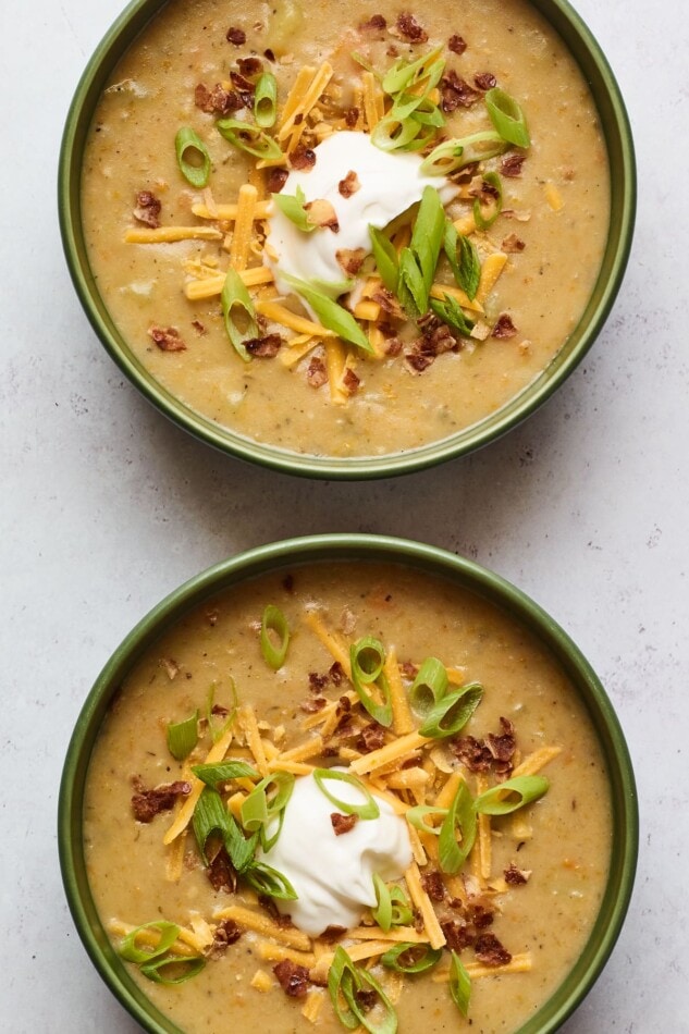 An overhead shot of two bowls of vegan potato soup. Both bowls have been topped with vegan sour cream, vegan shredded cheese, "bacon" bits, and chopped green onions.