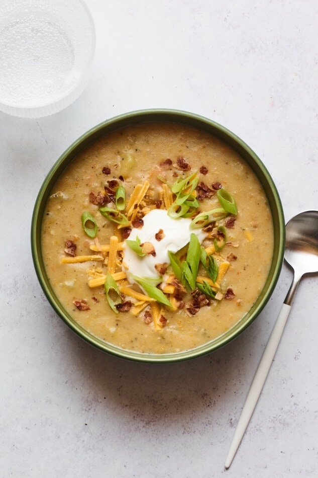 A bowl of vegan potato soup topped with vegan sour cream, vegan shredded cheese, "bacon" bits, and chopped green onions. A silver spoon rests aside the bowl.