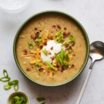 A bowl of vegan potato soup topped with vegan sour cream, vegan shredded cheese, "bacon" bits, and chopped green onions. A silver spoon rests aside the bowl.