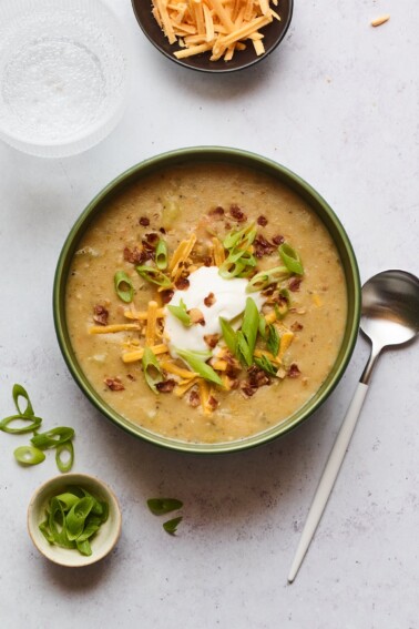 A bowl of vegan potato soup topped with vegan sour cream, vegan shredded cheese, "bacon" bits, and chopped green onions. A silver spoon rests aside the bowl.