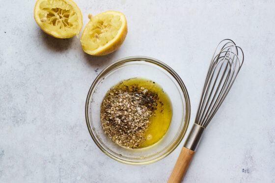 Spices and oil in a glass bowl. A whisk is resting next to the bowl as well as two lemon halves that have been juiced.