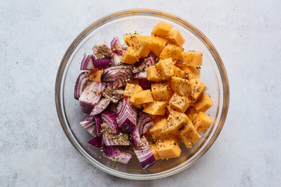 A glass bowl filled with red onion and butternut squash sprinkled with spices.
