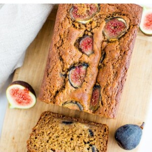 Loaf of fig bread on a wood cutting board with a few slices cut. Bread is surrounded by fresh figs.