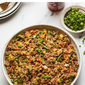 Serving bowl with quinoa fried "rice". Serving spoon is to the side. Sriracha bottle and a bow of chopped green onions is also to the side.