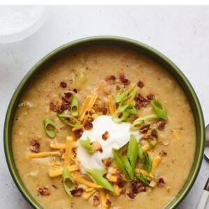 A bowl of vegan potato soup topped with vegan sour cream, vegan shredded cheese, "bacon" bits, and chopped green onions. A silver spoon rests aside the bowl.