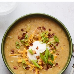 A bowl of vegan potato soup topped with vegan sour cream, vegan shredded cheese, "bacon" bits, and chopped green onions. A silver spoon rests aside the bowl.