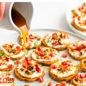 A hand is holding a small carafe of honey, drizzling the honey across a plate of sweet potato bites.