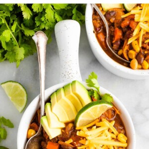 An overhead shot of a bowl of vegetarian chili, topped with avocado, shredded cheese, and a lime wedge.