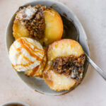 An overhead shot of a baked apple sliced in half with a scoop of ice cream and a caramel drizzle in a shallow bowl with a silver spoon.