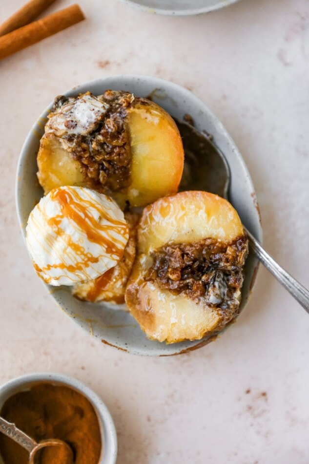 An overhead shot of a baked apple sliced in half with a scoop of ice cream and a caramel drizzle in a shallow bowl with a silver spoon.