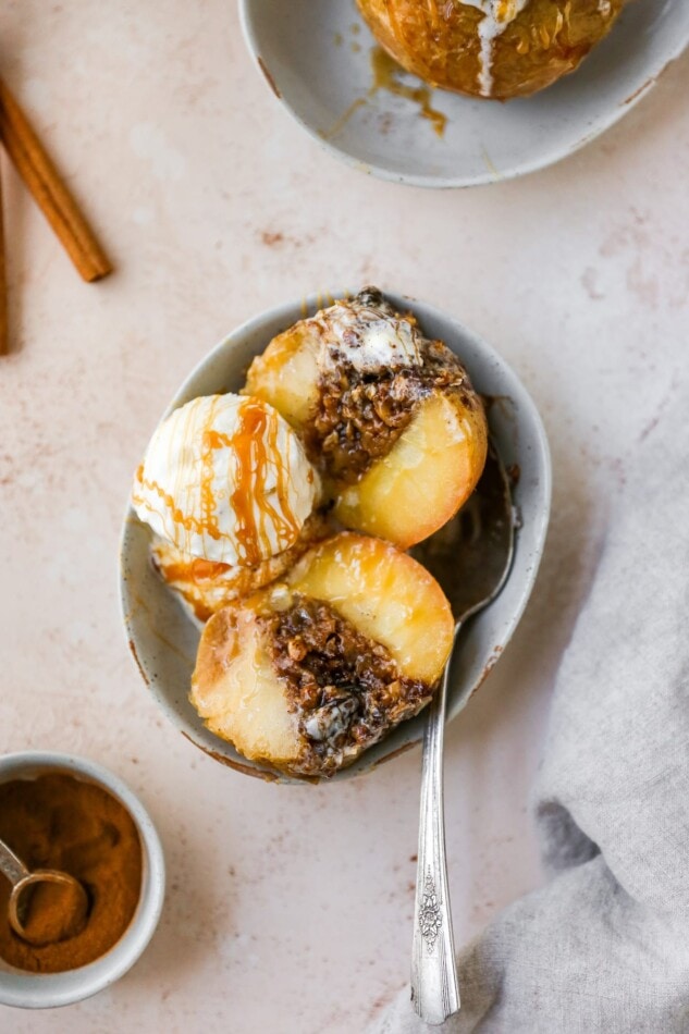 An overhead shot of a baked apple sliced in half with a scoop of ice cream and a caramel drizzle in a shallow bowl with a silver spoon.