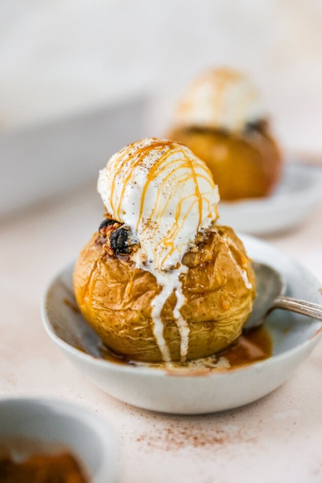 A baked apple with a scoop of ice cream and a drizzle of caramel on top, served in a small dish with a silver spoon.