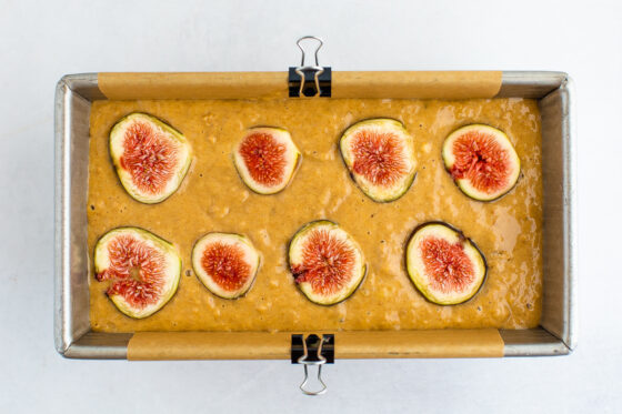 A bread pan filled with fig bread batter. 8 slices of fig are laid out on top of the bread.