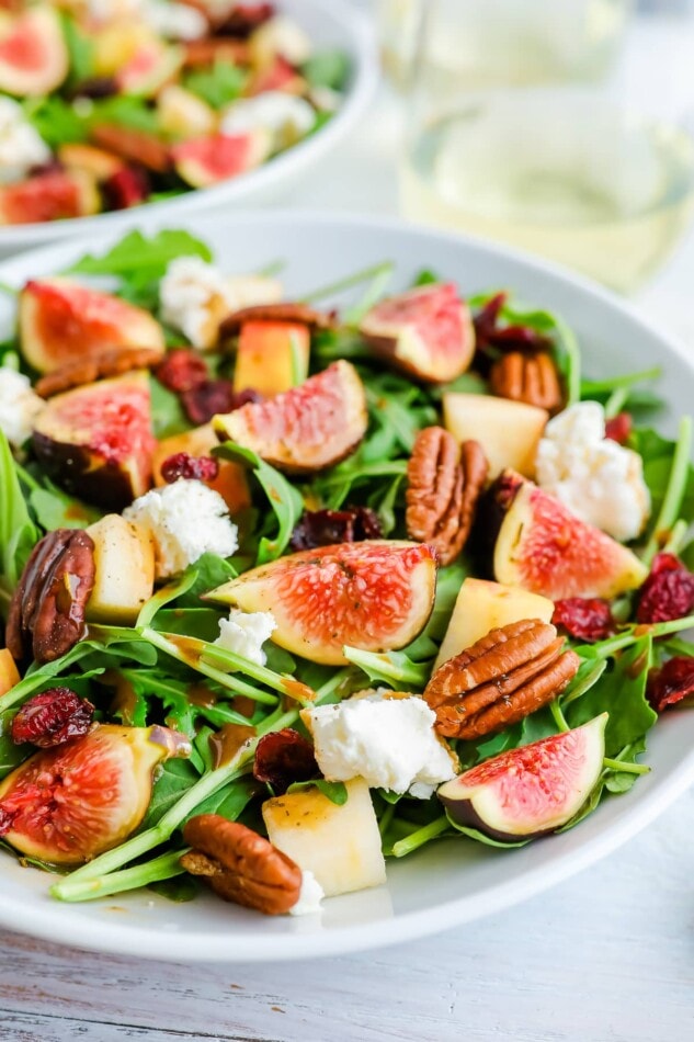 A bowl of fig salad with arugula and goat cheese. There is another bowl out of focus in the background alongside two stemless glasses of white wine.