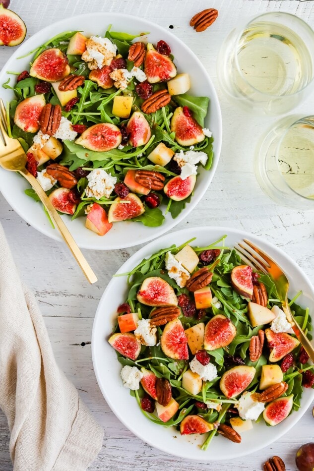 An overhead shot of two bowls of fig salad next to two glasses of white wine. A gold fork rests inside each bowl.