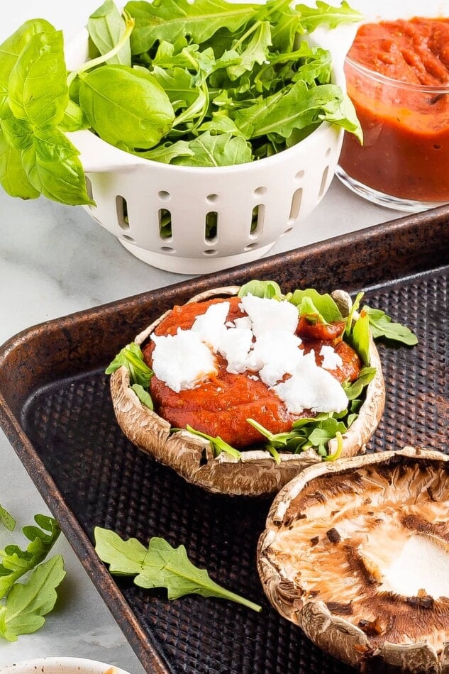 A portobello mushroom cap on a sheet pan, topped with arugula, pizza sauce, and goat cheese. Additional ingredients surround the sheet pan and there is another portobello mushroom cap with no toppings on the sheet pan.