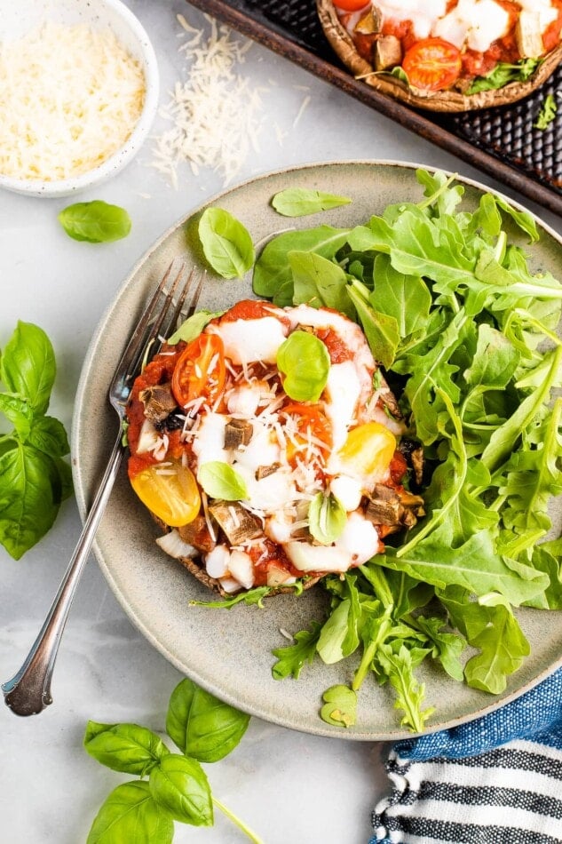 Overhead shot of a portobello mushroom pizza plated with arugula. A ramekin of cheese is in the top left corner and a silver fork rests on the plate.