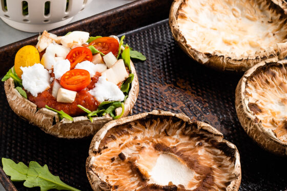 A portobello mushroom cap on a sheet pan, topped with arugula, pizza sauce, goat cheese, and sliced cherry tomatoes. Additional ingredients surround the sheet pan and there is another portobello mushroom cap with no toppings on the sheet pan.