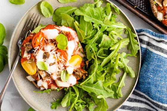 Overhead shot of a portobello mushroom pizza plated with arugula. A ramekin of cheese is in the top left corner and a silver fork rests on the plate.
