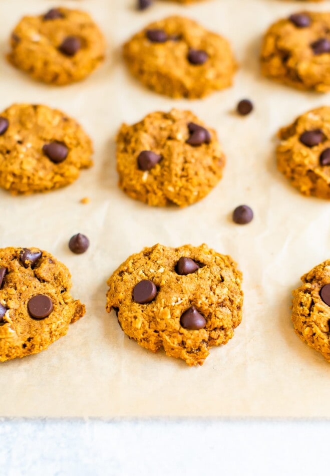 A piece of parchment paper with 9 cookies neatly in rows. There are extra chocolate chips sprinkled around the cookies.