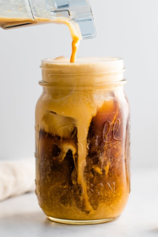 A mason jar with cold brew coffee and pumpkin cream being poured in to the top. The cream is mixing in with the cold brew.