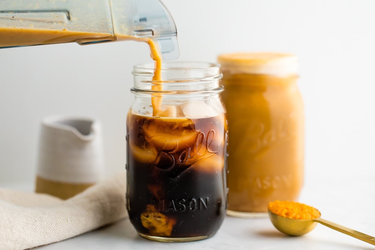 Pumpkin cream is being poured into cold brew and ice in a mason jar. A full mason jar with pumpkin cream cold brew is in the background
