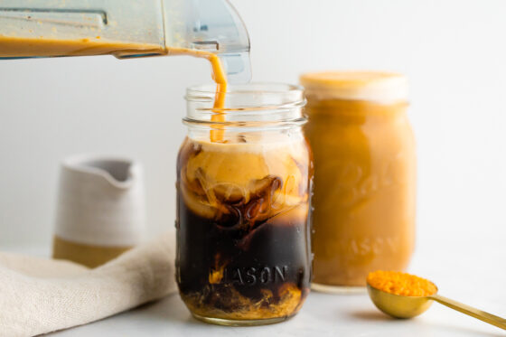 Pumpkin cream is being poured into cold brew and ice in a mason jar, creating a swirling affect as it pours in. A full mason jar with pumpkin cream cold brew is in the background