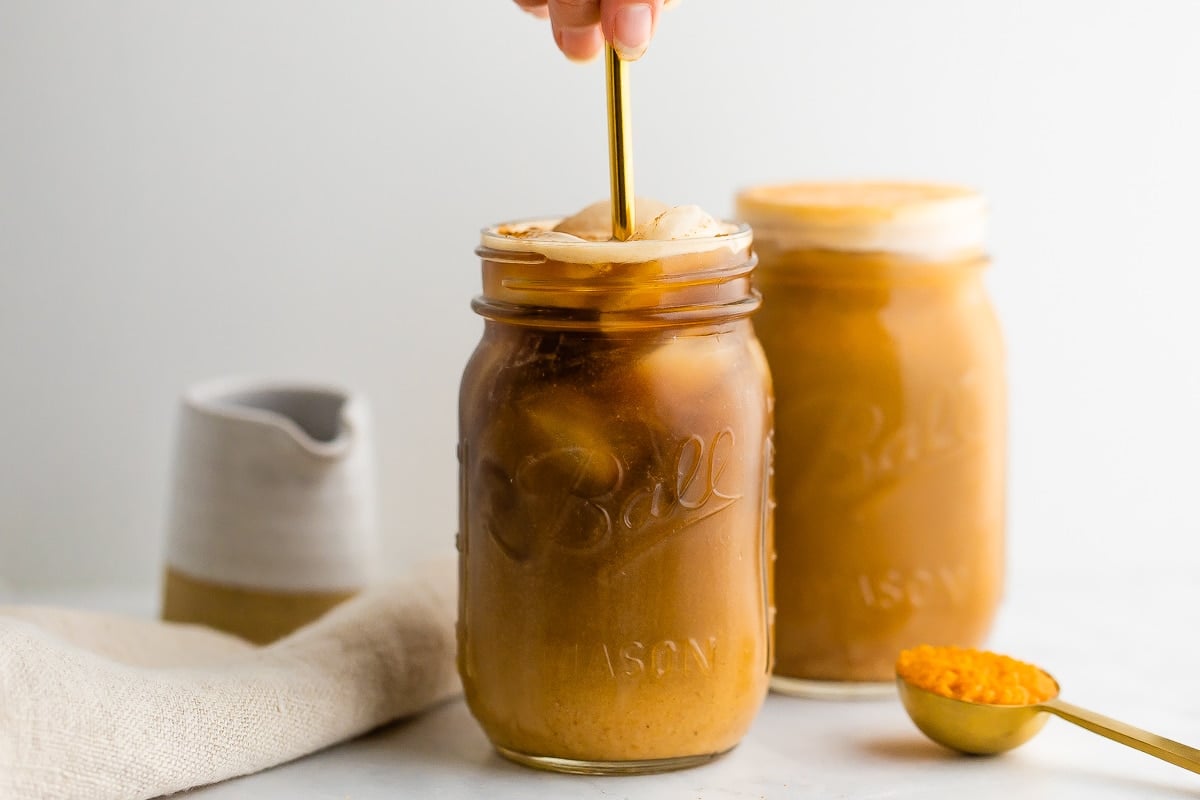 Pumpkin cream is being mixed into cold brew in a mason jar with a gold metal straw. A full mason jar with pumpkin cream cold brew is in the background.