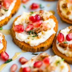 A sweet potato bite topped with pomegranate arils, pecans and fresh thyme is in focus, surrounded by other bites that are not in focus.