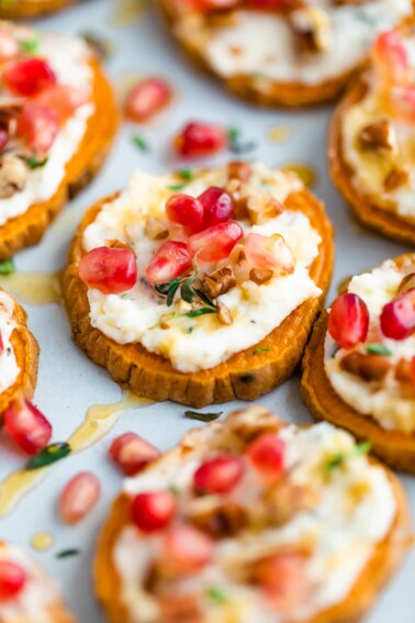 A sweet potato bite topped with pomegranate arils, pecans and fresh thyme is in focus, surrounded by other bites that are not in focus.